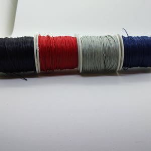 May include: A selection of waxed cord spools in black, red, grey, and navy blue. The cord is neatly wound on white plastic spools. The image is set against a white backdrop.