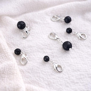 May include: Several small, silver-colored lobster clasp charms with black, porous lava rock beads. The charms are displayed on a white, textured fabric background. The beads are round and have a rough texture.