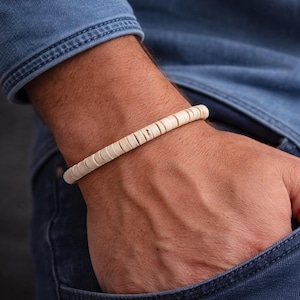 Può includere: Un braccialetto di perline color crema è indossato al polso. Il braccialetto è composto da piccole perline rotonde infilate insieme. La persona indossa una camicia di jeans e jeans. Il braccialetto è un accessorio neutro.
