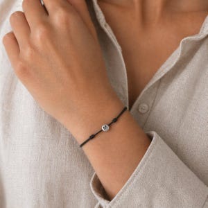 May include: A black cord bracelet with a small, round, silver charm bearing the letter "M". The bracelet is knotted on either side of the charm and is worn on a wrist.