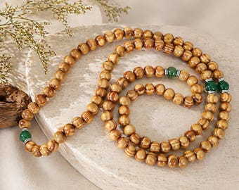May include: A wooden bead necklace with a natural wood grain pattern. The necklace features round beads and is accented with green gemstone beads. The beads are strung together, creating a layered effect.