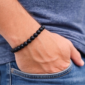 May include: A black beaded bracelet is worn on a wrist. The bracelet is made of matte black beads. The person is wearing a blue t-shirt and blue jeans. The person's hand is in their jeans pocket.
