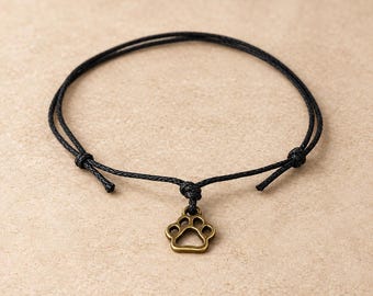 Puede incluir: Una pulsera de cordón negro con un dije de metal en tono bronce con forma de huella de animal. La pulsera está hecha de un cordón negro trenzado fino con nudos deslizantes ajustables a cada lado para asegurar el ajuste en la muñeca.