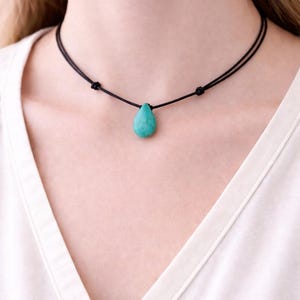 May include: A turquoise teardrop pendant necklace. The pendant hangs from a black cord choker. The necklace is worn against a white shirt.