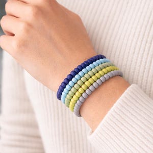 Op de afbeelding: Een stapel van zes kleurrijke spiraalarmbanden in blauw, groen en grijs op een pols. De armbanden zijn gemaakt van een flexibel materiaal. Op de achtergrond is een witte trui te zien. Elke armband is ongeveer 0,5 cm dik.