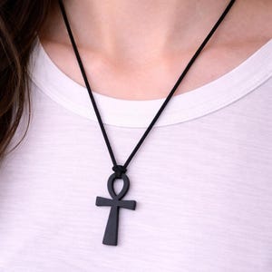 Könnte beinhalten: Schwarze Ankh-Anhänger-Halskette. Der Anhänger ist dunkel und einfarbig, an einer schwarzen Kordel befestigt. Das Ankh-Symbol ist ein Kreuz mit einer Schlaufe oben. Die Halskette wird über einem weißen Hemd getragen.