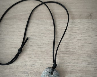 May include: A gray stone pendant necklace with a black cord. The oval-shaped stone has a natural, textured appearance. A US quarter-sized coin is next to the pendant for scale. The necklace is displayed on a light wood surface.