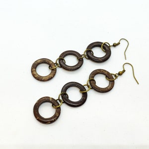 Może przedstawiać: Para brązowych kolczyków z muszli kokosa z pozłacanymi metalowymi pierścieniami łączącymi. Kolczyki mają prosty i elegancki design.