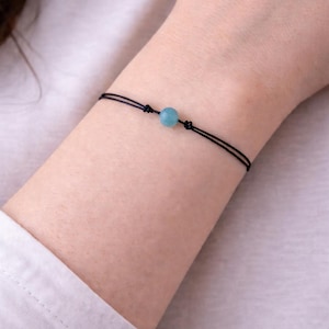 May include: A minimalist bracelet featuring a light blue bead centered on a black cord. The cord is knotted on each side of the bead. The bracelet is worn on a wrist, against a white background.