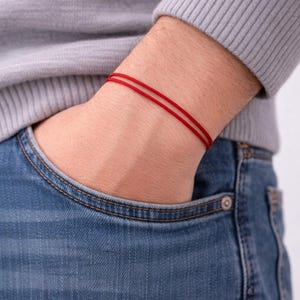 Puede incluir: Primer plano de una pulsera de hilo rojo en la muñeca. La pulsera está hecha de dos hebras paralelas de hilo rojo. La persona lleva un jersey gris y vaqueros azules.