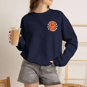 May include: A navy blue crewneck sweatshirt featuring a graphic on the left chest. The graphic consists of an orange smiley face surrounded by small white baseballs and the text "DETROIT TIGERS" in orange lettering arranged in a circular pattern.