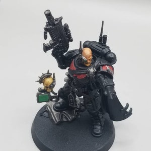 May include: A miniature figurine of a warrior in black armor with red accents, holding a black rifle. The figure has a bald head, a cybernetic eye, and a skull-shaped accessory. It stands on a black circular base.