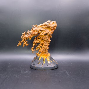 May include: A hand-painted miniature volcano erupting with orange and yellow lava. The base is dark gray with yellow highlights, and the lava flow has a textured, porous appearance. This fantasy terrain piece is suitable for tabletop gaming.