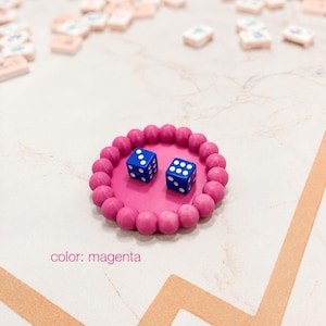 Puede incluir: Una bandeja circular de color magenta con un borde de cuentas que contiene dos dados azules con puntos blancos. La bandeja presenta un centro plano y liso y un borde esférico texturizado. El texto color: magenta aparece en la esquina inferior.