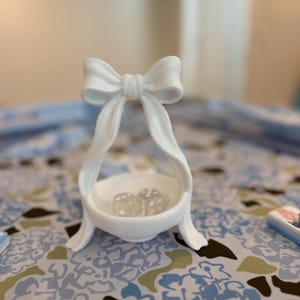 May include: A white ceramic bowl featuring a decorative bow handle that arches over the centre. The bowl contains two small, translucent dice. The object has a smooth, matte finish and stands on a base with two curved, ribbon-like supports.