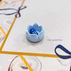 Pode incluir: Um suporte em forma de lótus azul pastel contendo um pequeno dado de seis faces azul escuro. O suporte apresenta pétalas arredondadas que formam uma estrutura em forma de taça, e o texto color: pastel blue está impresso à direita.