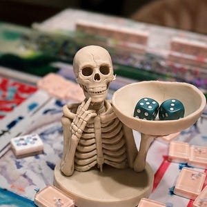 May include: A beige skeleton figurine with a skull and ribcage, posed with one hand near its chin and the other holding a shallow bowl. Inside the bowl are two teal six-sided dice with white pips. The figure stands on a circular base.