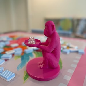 Könnte beinhalten: Eine leuchtend pinke Affenfigur in sitzender Position auf einem runden Sockel, die ein kleines rundes Tablett in den Händen hält. Ein einzelner, durchsichtiger, sechsseitiger Würfel mit weißen Punkten liegt auf dem Tablett der Affenskulptur.