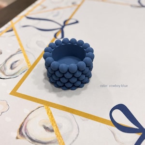 Puede incluir: Un portavelas de color azul vaquero con forma cilíndrica cubierto por un patrón texturizado de pequeñas esferas uniformes. El objeto tiene un acabado mate liso y un centro hueco, con el texto color: cowboy blue impreso cerca.