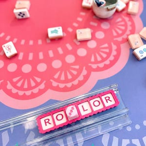 May include: A clear acrylic tile rack holding five white tiles with red letters spelling R O L O R, with a pink scalloped border. Scattered around are various game tiles and a small ceramic cup on a pink and purple patterned surface.