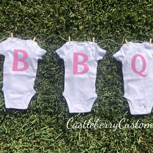 May include: Three white baby onesies with pink letters spelling out "BBQ" hanging on a clothesline.