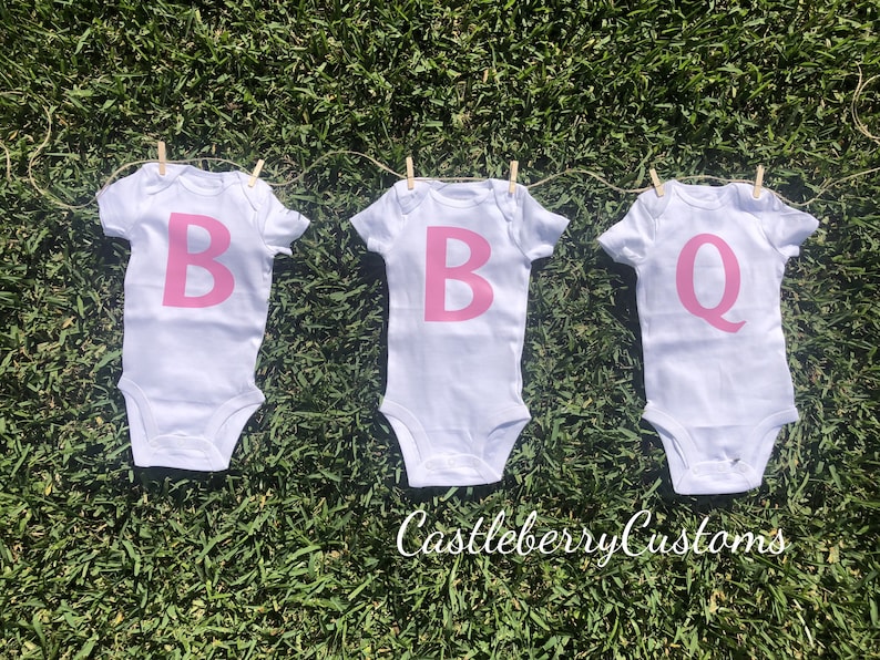 May include: Three white baby onesies with pink letters spelling out "BBQ" hanging on a clothesline.