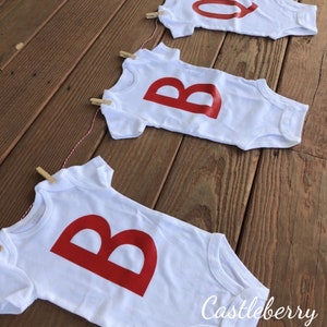 May include: A banner made of three white baby onesies with red letters spelling "BOO" hanging on a string with clothespins. The onesies are on a wooden surface. The text "Castleberry Customs" is visible in the bottom right corner of the image.