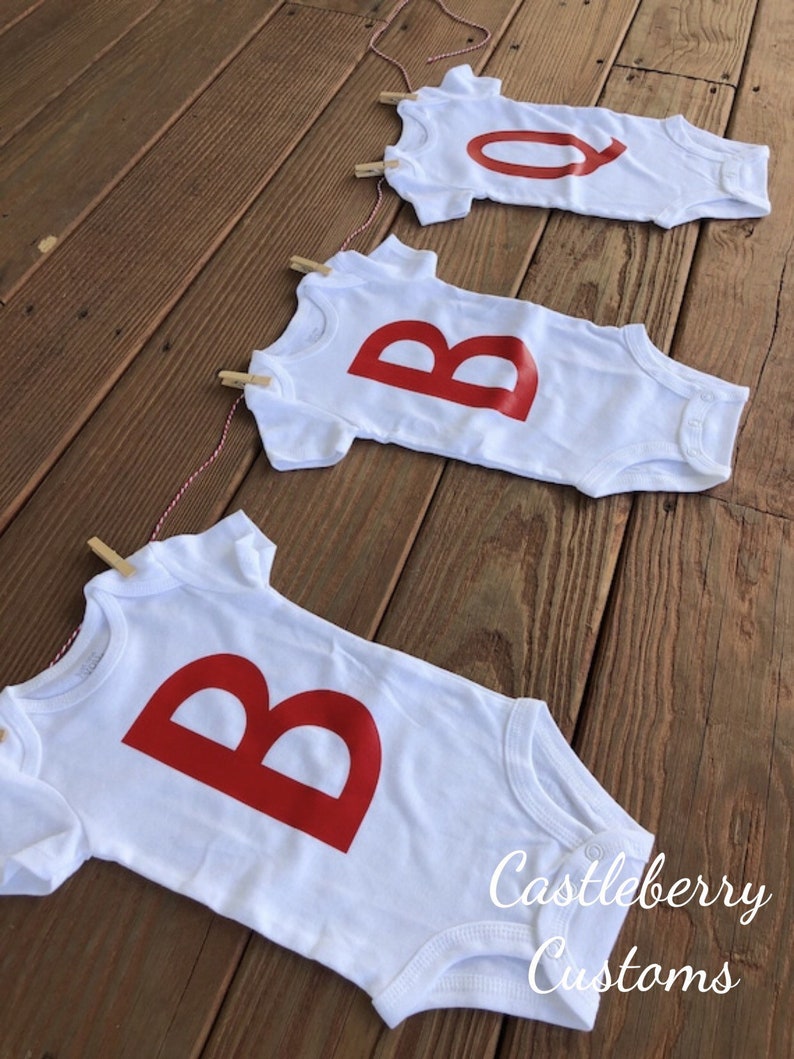 May include: A banner made of three white baby onesies with red letters spelling "BOO" hanging on a string with clothespins. The onesies are on a wooden surface. The text "Castleberry Customs" is visible in the bottom right corner of the image.