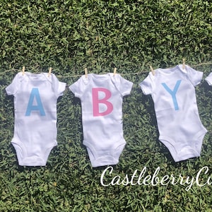 May include: Five white baby onesies with the letters "BABY" spelled out in pink and blue. The onesies are hanging on a clothesline with clothespins. The text "Castleberry Customs" is visible at the bottom of the image.