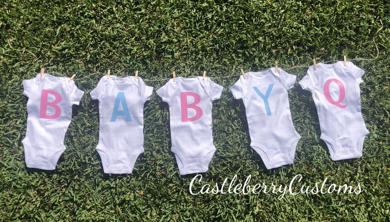 May include: Five white baby onesies with the letters "BABY" spelled out in pink and blue. The onesies are hanging on a clothesline with clothespins. The text "Castleberry Customs" is visible at the bottom of the image.