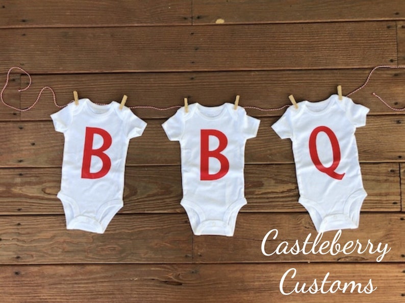 May include: Three white baby onesies with red letters spelling out "BBQ" hanging on a clothesline with red string and wooden clothespins. The onesies are on a wooden background. The text "Castleberry Customs" is written below the onesies.