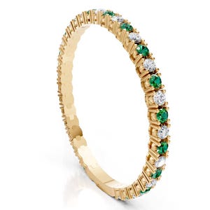 May include: A gold band ring featuring alternating round-cut emerald and diamond gemstones. The ring's setting is a delicate, detailed design, showcasing the gemstones' brilliance. The ring is set against a white background.