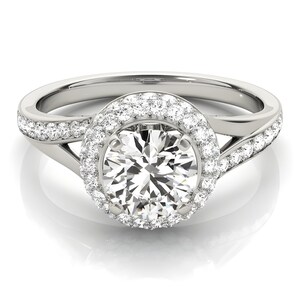 May include: A silver engagement ring with a large, round, clear center stone surrounded by smaller stones. The band features a split design with additional small stones. The ring is set against a white background.