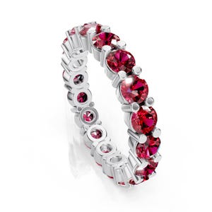 May include: A silver eternity ring featuring a continuous row of vibrant, round-cut, red gemstones. The gemstones are set closely together, creating a full circle of color. The ring's metal band is polished, reflecting light.