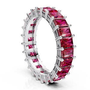 May include: A silver eternity ring featuring a continuous row of square-cut, vibrant pink gemstones. The ring's setting has a detailed, geometric design, with small metal bars securing each gemstone. The gemstones are a deep, saturated pink color.