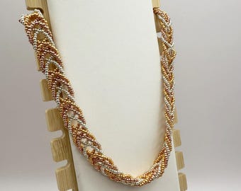 May include: A braided necklace displayed on a white stand. The necklace is made of small beads in gold, silver, and copper colors, creating a textured, interwoven design. The stand is made of light-colored wood.