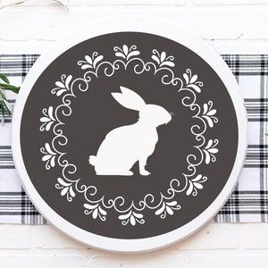 May include: A round wooden sign with a white silhouette of a rabbit inside a white floral wreath on a dark gray background.