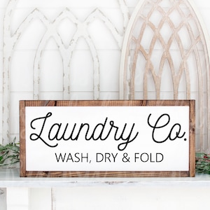 Op de afbeelding: Een houten bord met een witte achtergrond en een bruine lijst. Het bord heeft de tekst "Laundry Co. WASH, DRY & FOLD" in zwarte, cursieve letters.
