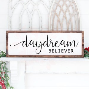 May include: A wooden sign with a brown frame and a white background. The sign says "daydream believer" in black cursive lettering.