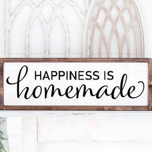 May include: A wooden sign with a white background and brown frame. The sign says "Happiness is homemade" in black cursive lettering.