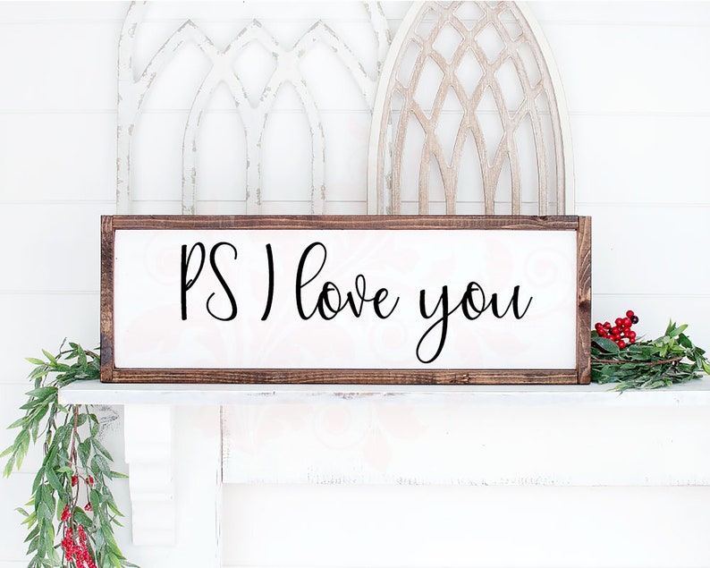 May include: A rectangular wooden sign with the phrase "PS I love you" in black script. The sign has a dark brown frame and a white background. Decorative greenery and berries are on the shelf below.