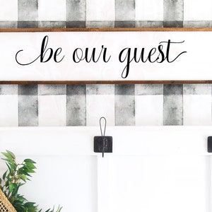 May include: A white wooden sign with a brown frame hanging on a black and white checkered wall. The sign says "be our guest" in black cursive lettering.