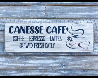 May include: A rustic wooden sign with the text "Canesse Cafe" in black lettering. The sign also includes the text "Coffee - Espresso - Lattes" and "Brewed Fresh Daily". A stylized image of a coffee cup with steam and a heart is on the right side of the sign.