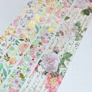 May include: Decorative washi tape rolls with floral and botanical designs. The tapes feature various patterns, including gold foil accents, roses, and script. The color palette includes pink, peach, green, and gold on a white background.