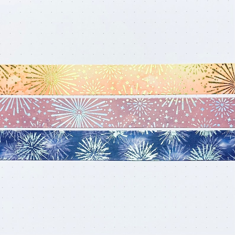 May include: Three rolls of washi tape with a firework pattern. The top roll is peach with gold glitter, the middle roll is pink with silver glitter, and the bottom roll is blue with silver glitter.