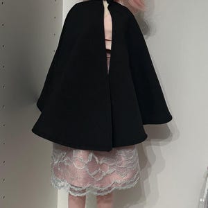 May include: A pink-haired doll wearing a black cape, a pink and white lace skirt, and pink shoes. The doll has elf ears and is standing in front of a white wall.