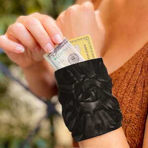 May include: A black arm band with a floral pattern. The arm band has a pocket that can hold cash and a credit card. The credit card is visible in the pocket and has the text "AMERICAN" on it. The arm band is worn on a person's arm.