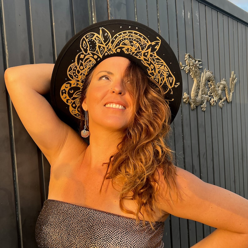 Puede incluir: Una mujer con el pelo largo, ondulado y casta&ntilde;o claro lleva un sombrero negro de ala ancha con dise&ntilde;os geom&eacute;tricos dorados. Lleva un top negro brillante sin tirantes. El sombrero est&aacute; ligeramente inclinado hacia un lado y la mujer mira hacia arriba.