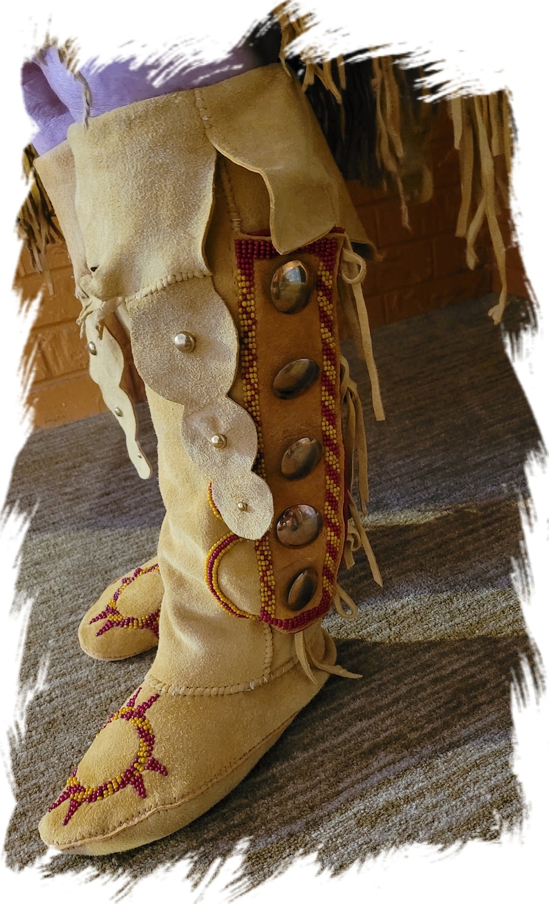May include: A pair of tan leather moccasins with intricate beadwork and decorative stitching. The moccasins have a red and yellow beaded design and are adorned with metal accents.