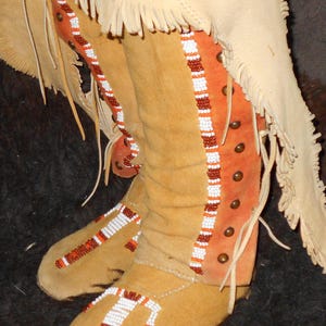May include: Close-up of a pair of tan leather moccasins with intricate beadwork. The moccasins have a fringe detail and are decorated with orange, white, and brown beads.